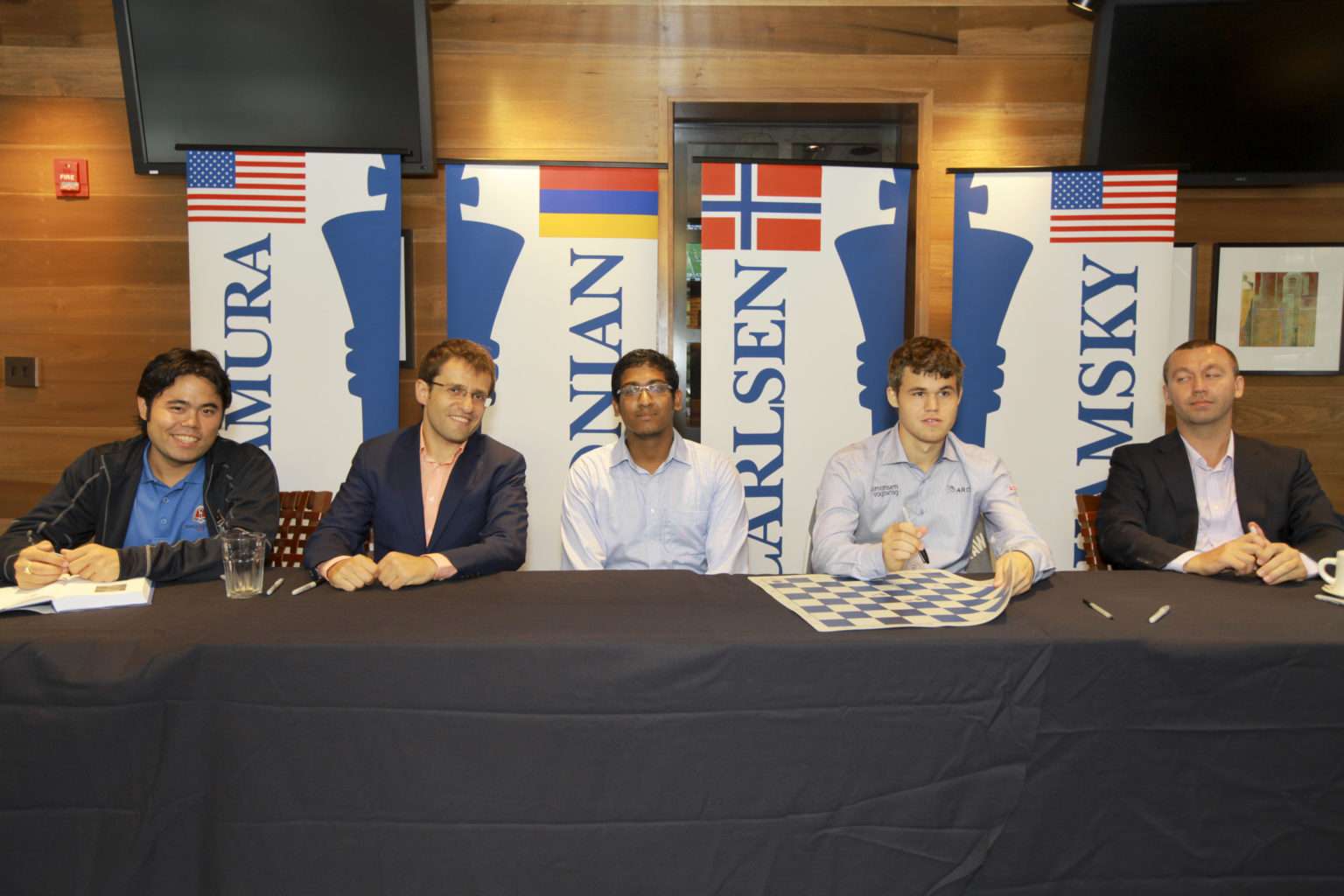 Five men sit at a table in front of chess-themed banners and flags, with a chessboard and papers on the table in a wood-paneled room.