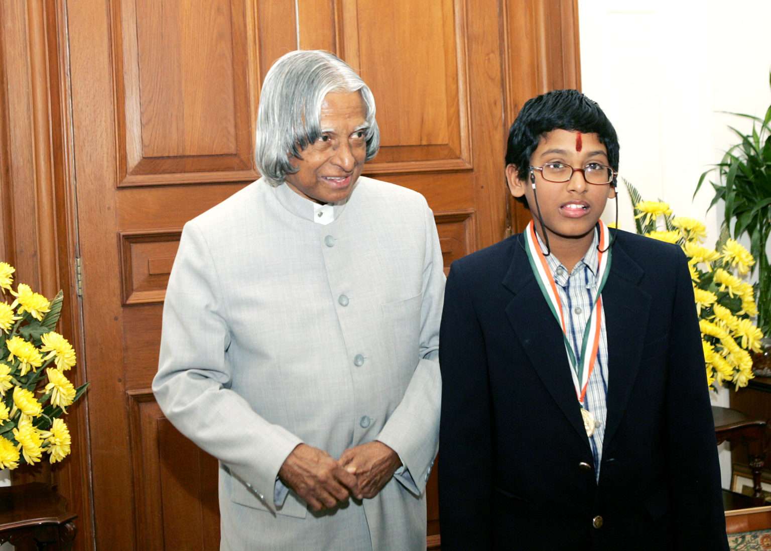 An older man in a light suit stands beside a young boy wearing glasses, a medal, and a striped shirt with a blazer. They are indoors with yellow flowers and wooden paneling in the background.